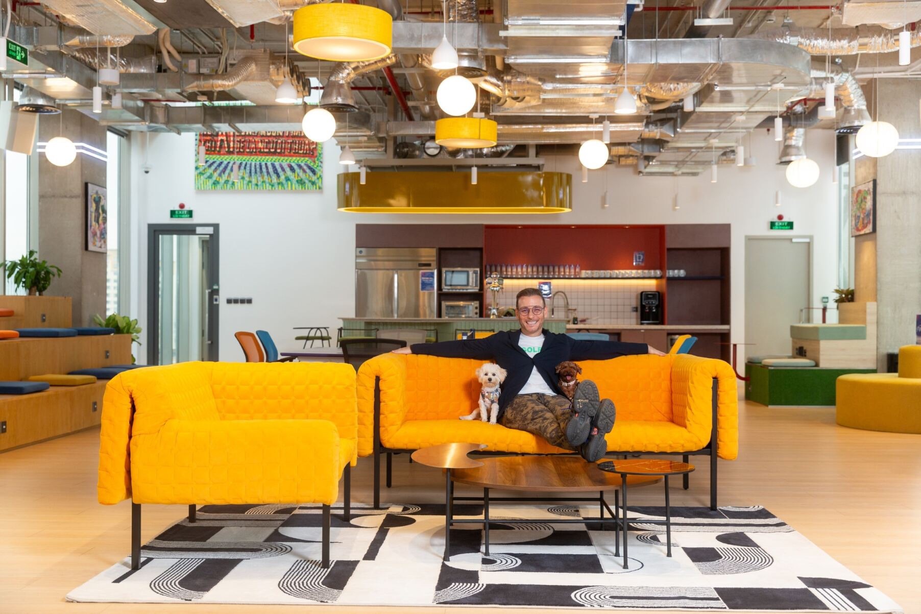 A man sitting on a bright orange couch with two small dogs in a modern, vibrant coworking space, illustrating the hybrid workplace model.