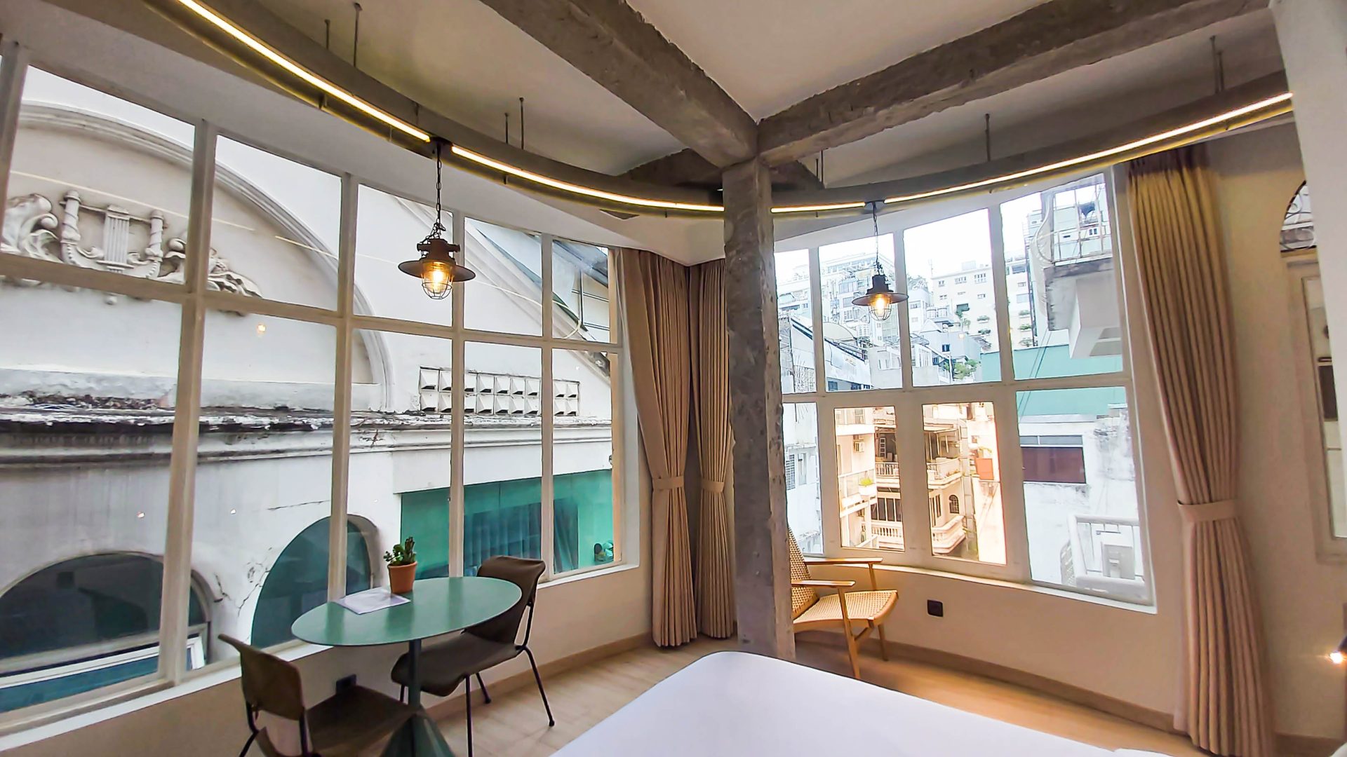 A well-lit room with large windows offering a view of an urban street, featuring a small round table with chairs, warm lighting, and neutral-toned curtains, providing a cozy and inspiring workspace for digital nomads.